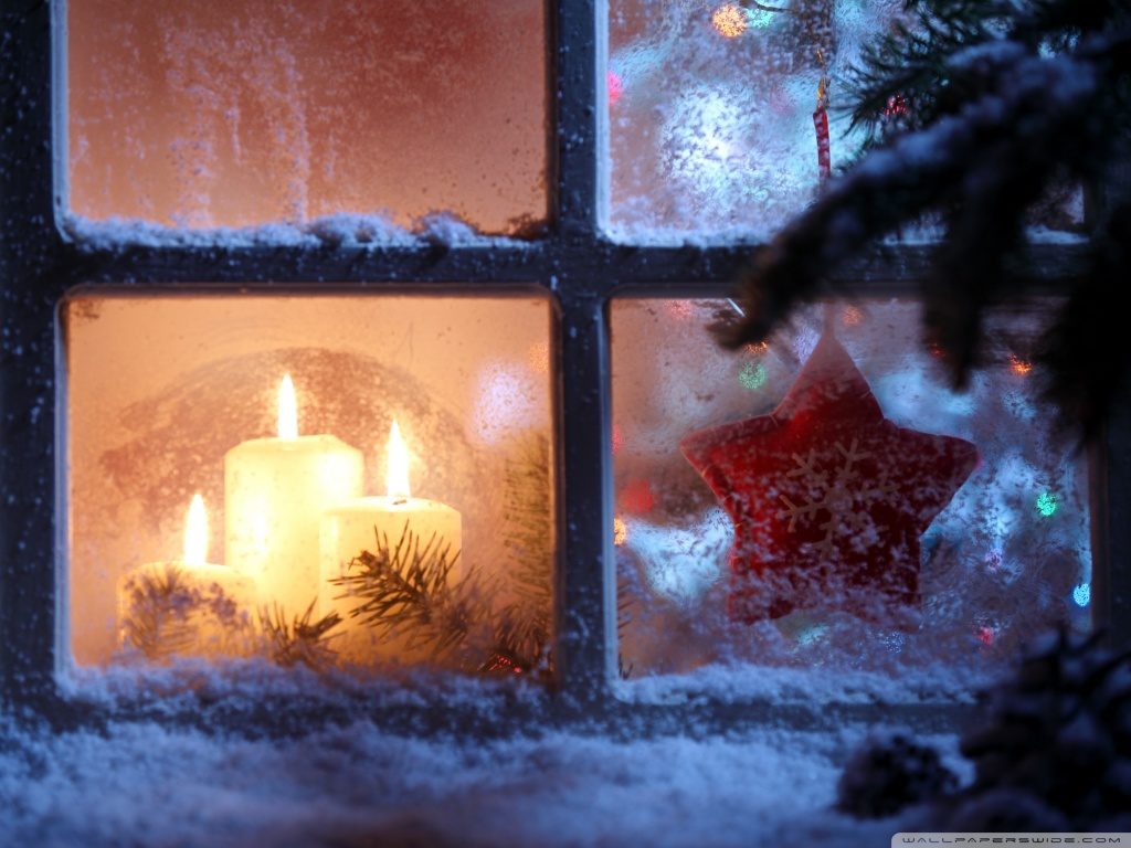 Pohled na zasněžené okno, za kterým jsou tři hořící bílé svíčky a červená ozdoba ve tvaru hvězdy.