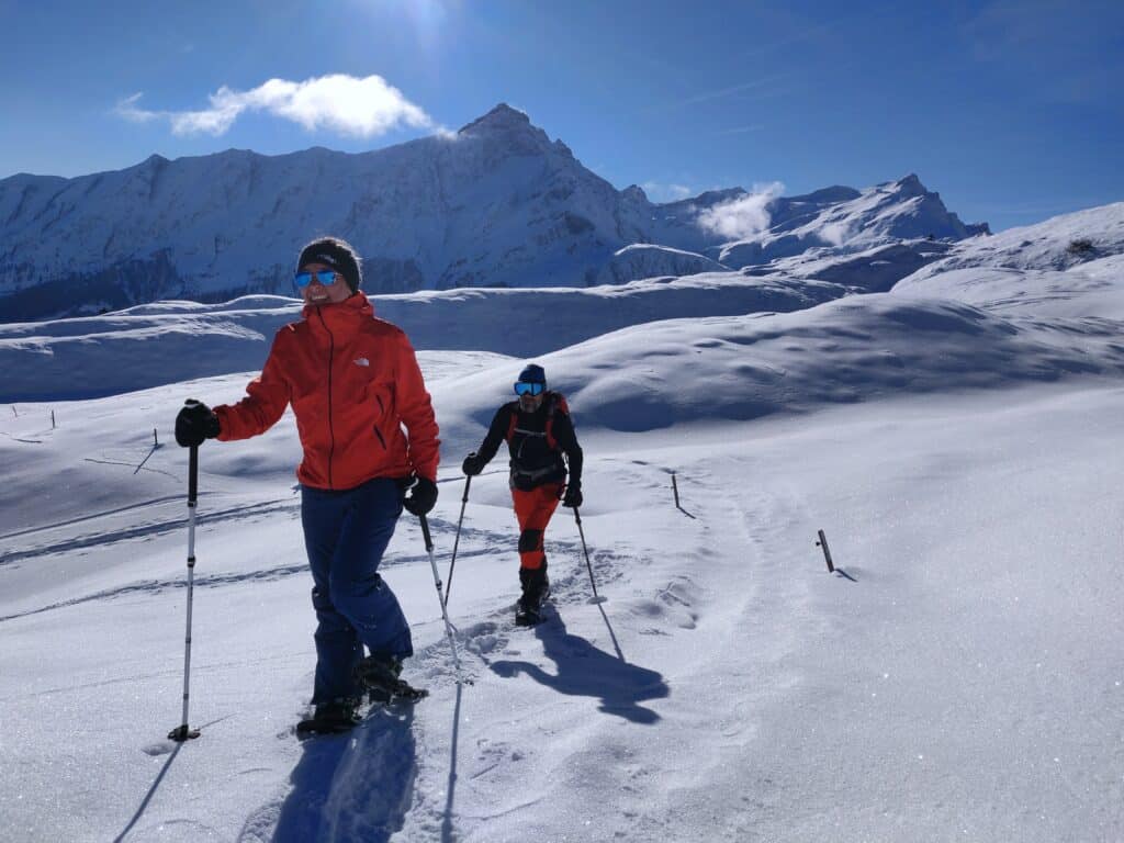 Žena a muž kráčí zasněženou krajinou pod vysokými horami ve sněžnicích.