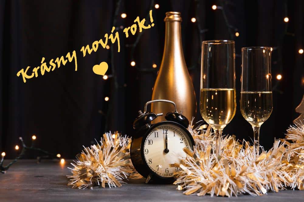 Novoroční zátiší v podobě dvou sklenic šampaňského, lahví, budíkem s ručičkami na dvanáctce a vánočním řetězem s přáním k novému roku.
