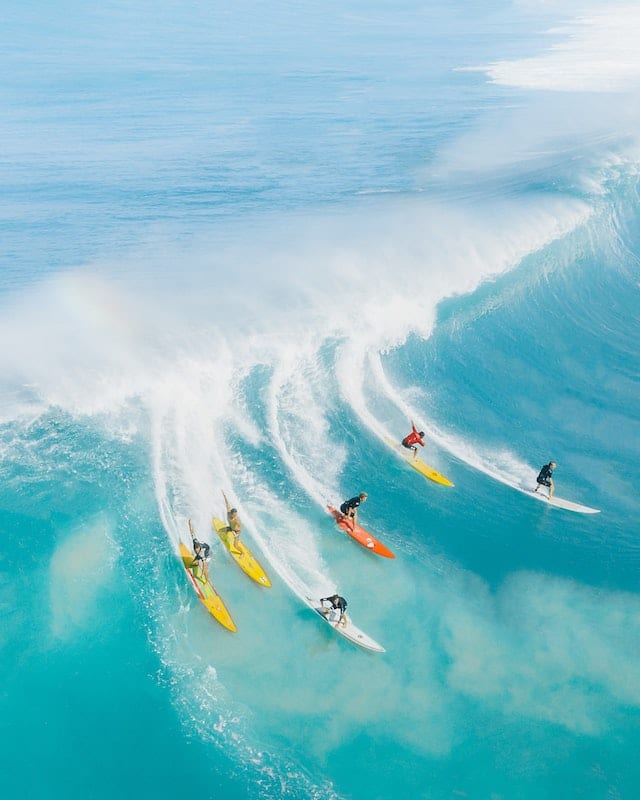 Průzračné modré moře na Havaji se zvedající se vlnou, na ní se sjíždí šest surfařů.